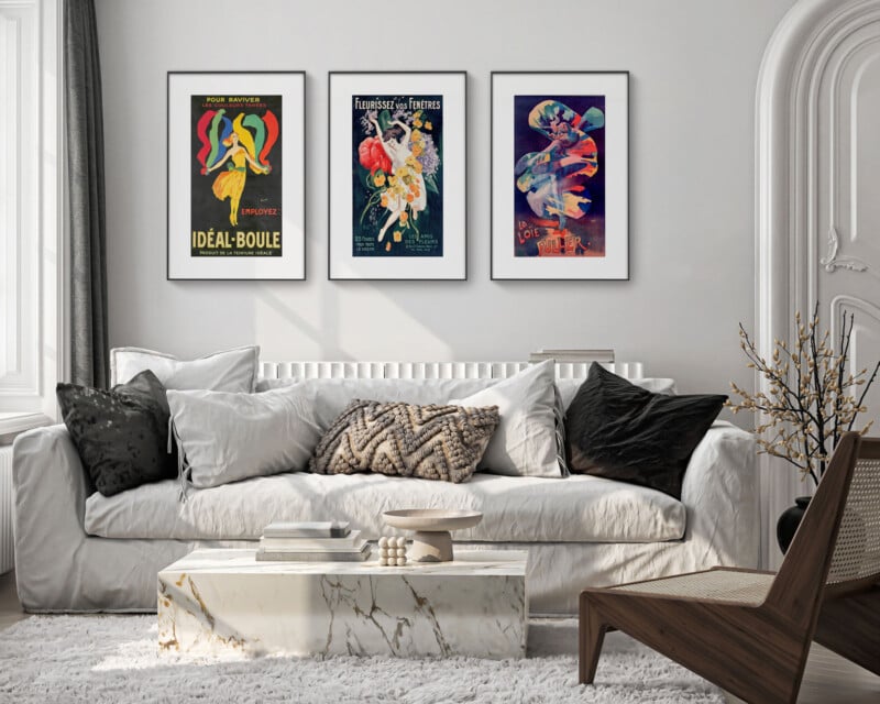 A modern living room with a white sofa and cushions, a marble coffee table, and framed vintage posters on the wall. The room features a neutral color palette and a minimalist decor with a woven chair and a small plant.