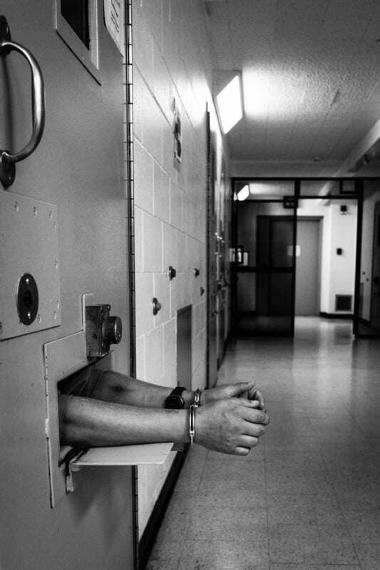 A monochromatic image of a prison corridor. Handcuffed hands extend through a small gap in a cell door to the left. The passageway is vacant, with closed doors and overhead fluorescent lighting.