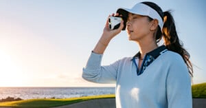 A person wearing a light blue sweater and white visor uses a golf rangefinder on a sunny day. A golf course and ocean are visible in the background.