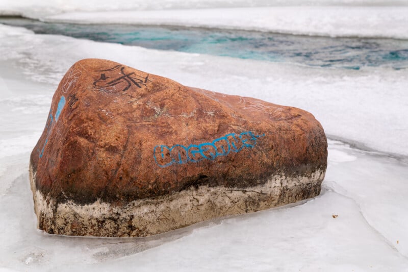 A large, weathered brown boulder sits on an icy surface with carved and painted graffiti. Patches of snow surround it, and a partially frozen blue stream flows nearby.