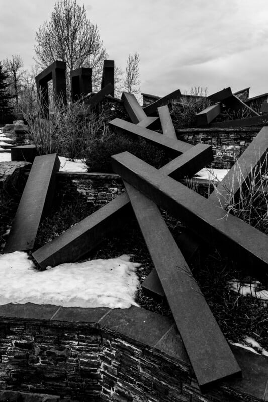 A black and white image of an outdoor abstract sculpture made of large, intersecting metal beams. Snow patches cover the ground, and bare trees are visible in the background.