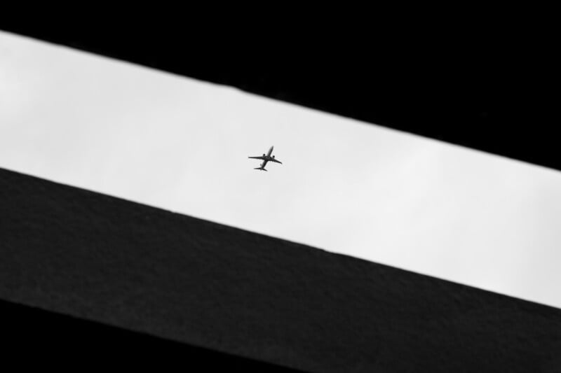 A silhouette of an airplane is visible through a narrow rectangular gap in a dark structure, set against a cloudy sky.