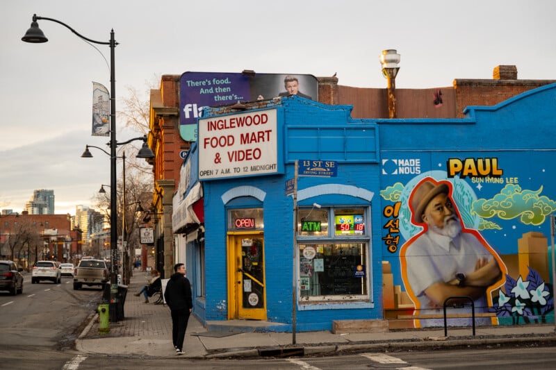 Street view of Inglewood Food Mart & Video with colorful mural of a man on the wall. The store has signs for ATM and is open. A person walks on the sidewalk, and a lamppost and buildings are in the background.