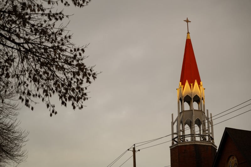 A church steeple with a red roof and a cross at its peak is illuminated against a gray, overcast sky. Bare tree branches are visible on the left side of the image.