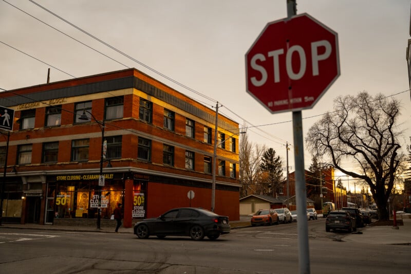 Street corner with a red "STOP" sign in the foreground and a brick building displaying "50% Closing Sale" signs. A black car is turning at the intersection, and a leafless tree stands by the sidewalk under an overcast sky.