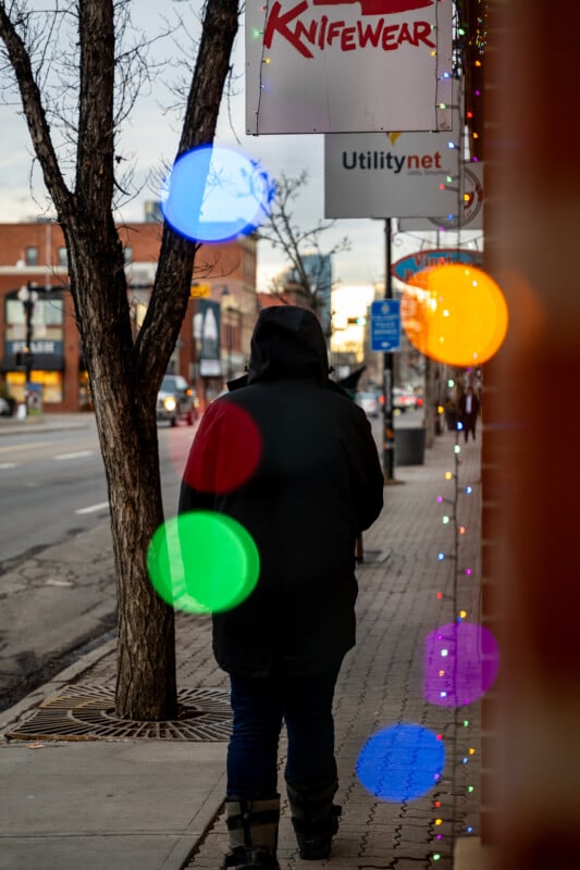 A person in a dark coat walks on a city sidewalk lined with holiday lights and colorful bokeh. The street is empty, with a leafless tree on the left and brick buildings in the background. Signs for "Knifewear" and "Utilitynet" are visible.