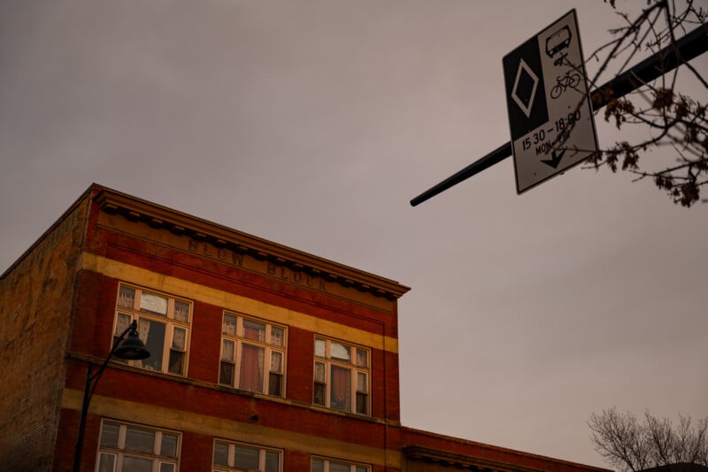 A red-brick building with "ELOW BLOCK" on the facade, under a cloudy sky. A street sign with a bus and bicycle symbol, along with time restrictions, appears in the foreground. Branches of a tree are visible on the side.