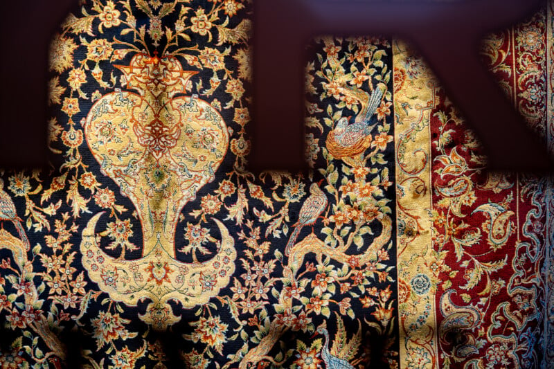 A richly detailed Persian carpet featuring intricate floral patterns in gold, red, and blue hues. The design includes symmetrical motifs and bird illustrations, creating an ornate and luxurious appearance.