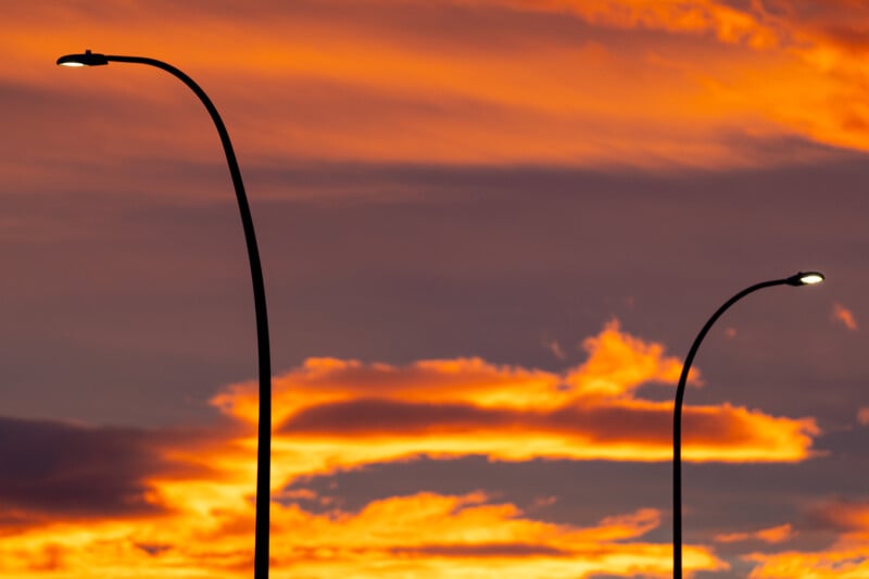 Two streetlights are silhouetted against a vibrant orange and purple sunset sky. The clouds create a dramatic backdrop of swirling colors, enhancing the contrast with the dark lamp posts in the foreground.