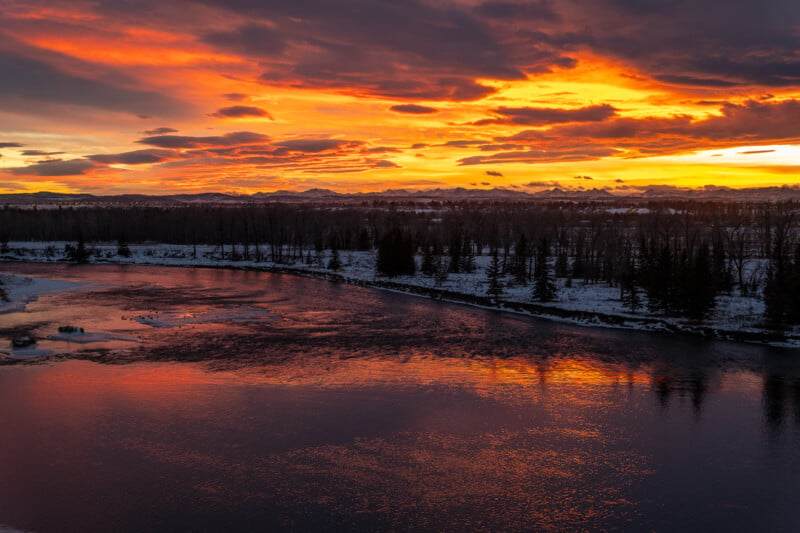 A vibrant sunset over a scenic river with orange and purple-hued clouds reflecting on the water. Snow-dusted trees line the riverbank, with a backdrop of distant mountains under the colorful sky.