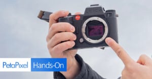 A person holding a Leica camera, pointing at the sensor with one finger. The image includes the PetaPixel logo and the words "Hands-On" at the bottom left corner. The camera is shown against a blurred gray background.