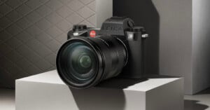 A Leica camera with a large lens is displayed on a geometric pedestal. The background features a textured, diamond-patterned surface in a neutral tone, casting soft shadows.