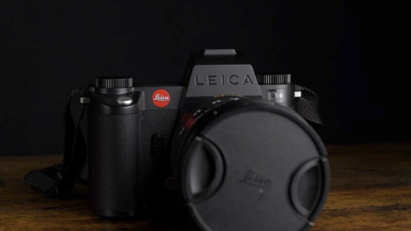 A black Leica camera with a lens cap on, prominently displaying the Leica logo, is placed on a wooden surface against a dark background.