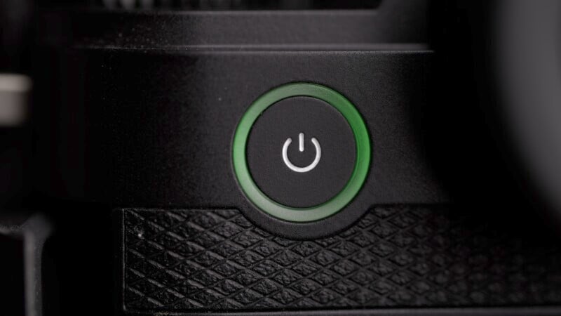 Close-up image of a black power button with a white power symbol, outlined by a green LED ring on a textured black surface.