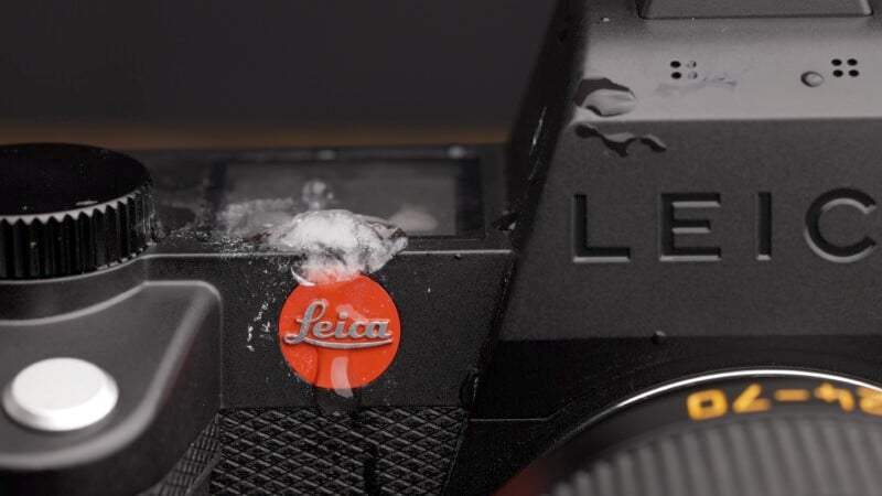 Close-up of a black Leica camera with a visible red Leica logo covered partially by snow. The camera body has textured grip and some visible snowflakes.