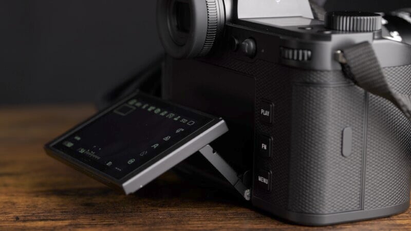 Close-up of a black digital camera on a wooden surface. The camera's tilt-out LCD screen is partially extended, displaying various settings icons, with the camera's textured grip visible. The background is dark and out of focus.