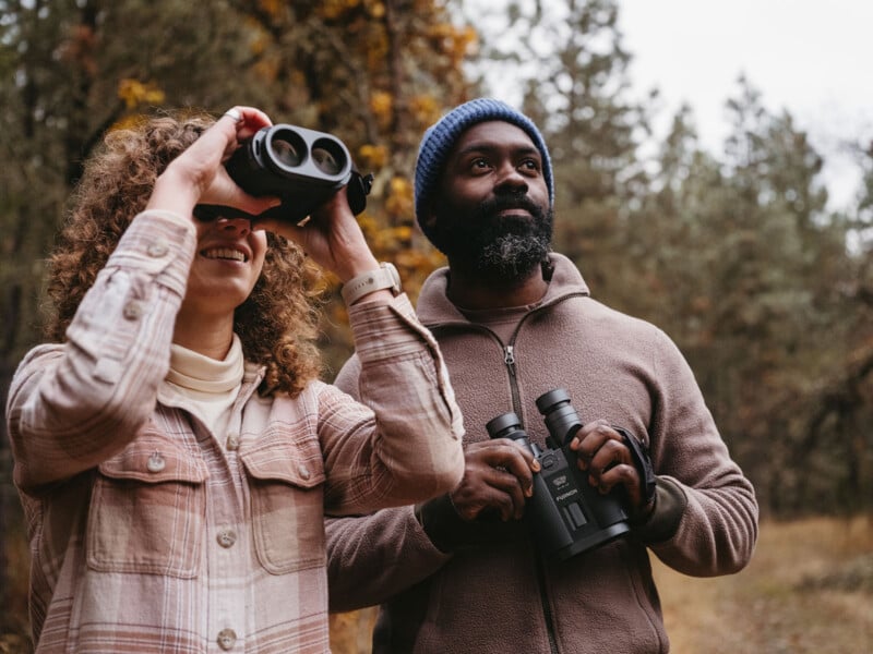 A man and woman are birdwatching in a forest. The woman, wearing a plaid jacket, looks through binoculars, while the man in a blue beanie and brown jacket holds his binoculars, gazing upward. The background features tall trees.