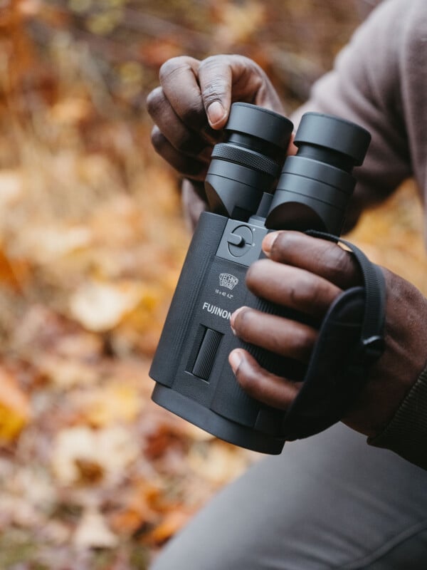 A person holding binoculars with both hands, surrounded by fallen autumn leaves on the ground. The individual is wearing a brown long-sleeved shirt, and the binoculars are branded with Fujinon.