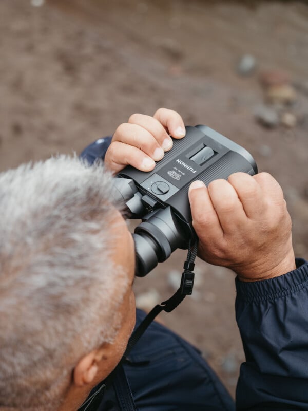 A person with short gray hair, seen from above, holds binoculars up to their eyes. They are wearing a dark jacket, and the background is a blurred, sandy terrain.