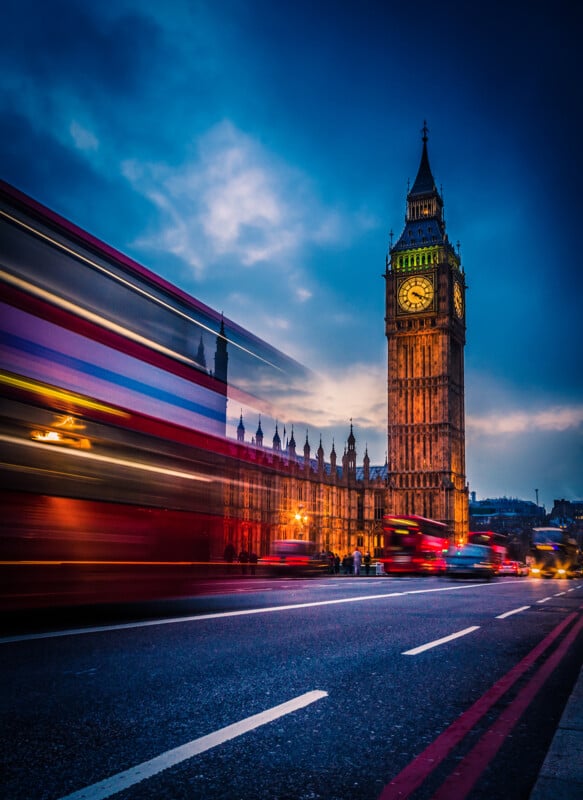 A blurred London double-decker bus speeds past in the foreground with Big Ben and the Houses of Parliament illuminated against a dusky blue sky. The scene captures the dynamic energy of London traffic and iconic architecture.