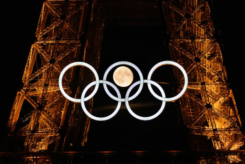 The picture depicts the Eiffel Tower illuminated in a warm golden hue at night. Large Olympic rings hang centrally, with the full moon perfectly positioned within the rings, generating a stunning visual impact.