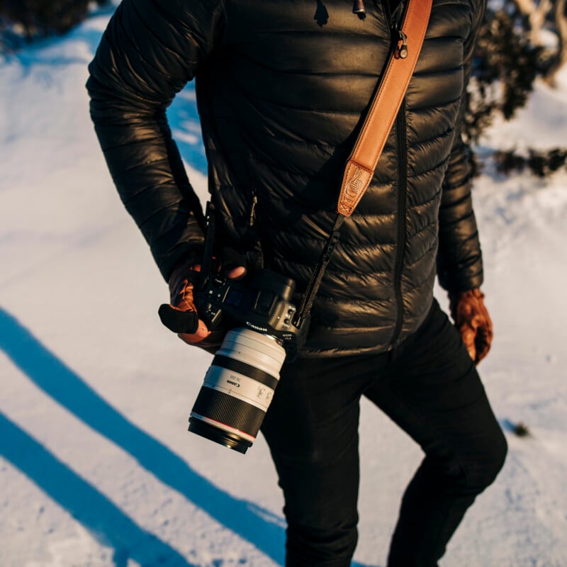 Person wearing a black puffer jacket and dark pants, holding a camera with a large lens. They are outdoors on a snowy landscape, with sunlight casting shadows on the ground. A leather camera strap is slung over their shoulder.