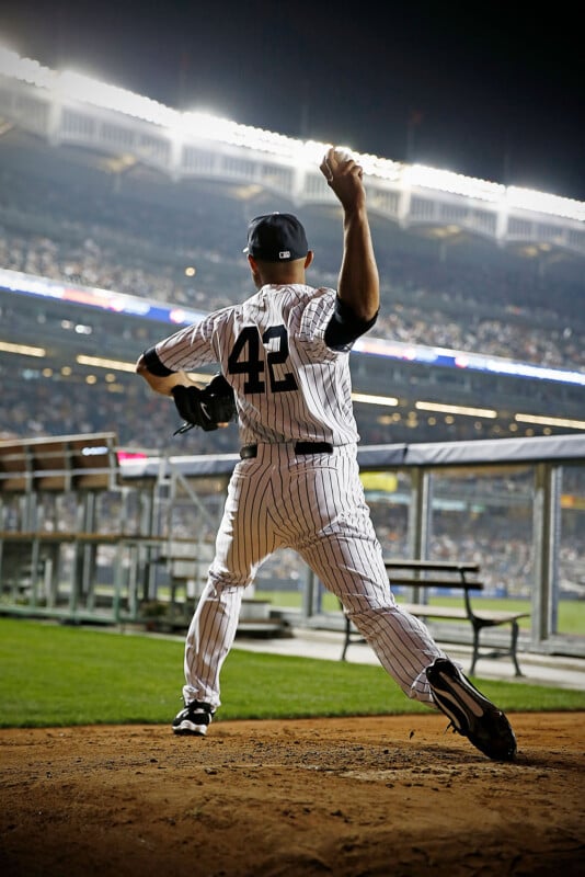 A baseball player wearing a jersey with the number 42 is warming up on the pitcher's mound. The stadium is packed with spectators and illuminated by bright lights, creating a dynamic, energetic atmosphere.