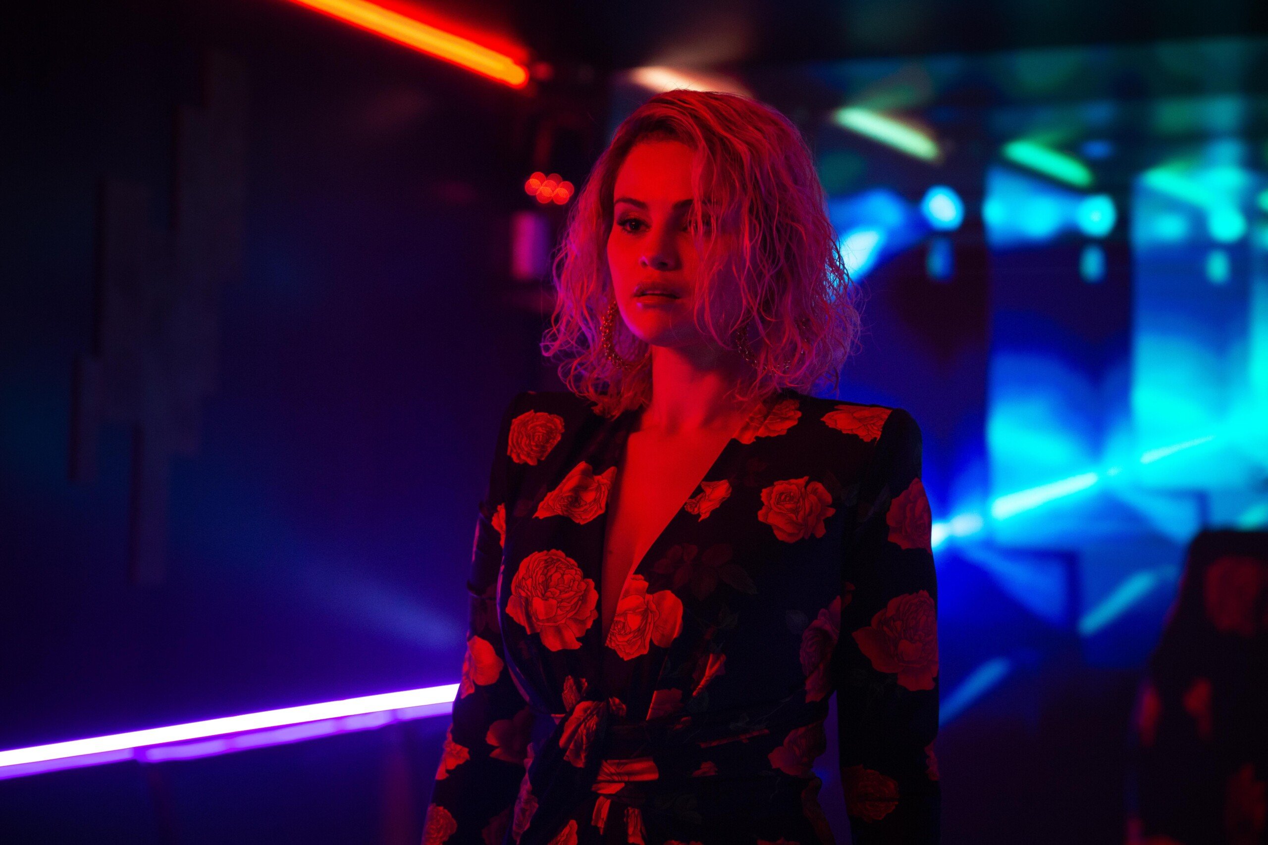 A person with curly hair stands in a dimly lit room, surrounded by neon red and blue lights. They wear a floral-patterned outfit. The ambient lighting creates a dramatic atmosphere.