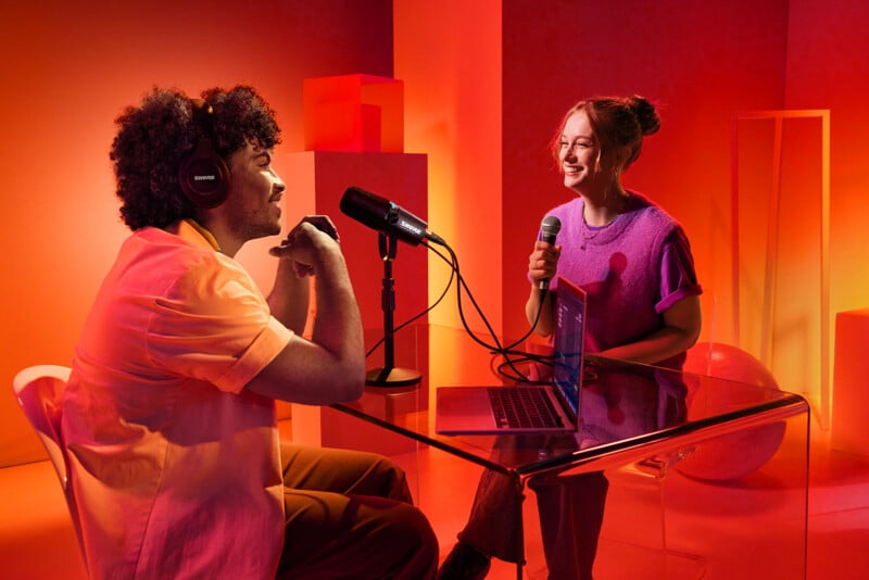 Two people sit at a glass table in a brightly lit studio, surrounded by red and orange decor. They are engaged in a podcast recording, each speaking into a microphone. A laptop is open on the table, and one person wears headphones.