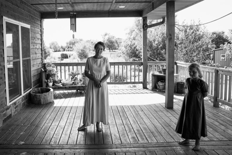 A monochrome image of a woman and a child standing on a wooden deck. The woman, donned in a light dress, stands centrally, while the child, dressed in darker attire, stands to the right. Sunlight casts prominent shadows on the porch. Trees are visible in the backdrop.