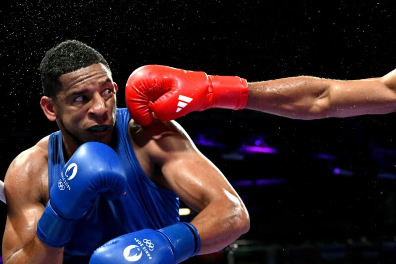 A boxer wearing blue braces for impact as an opponent's red-gloved fist approaches during a match. The background is dark, highlighting the intensity and focus of the athletes.