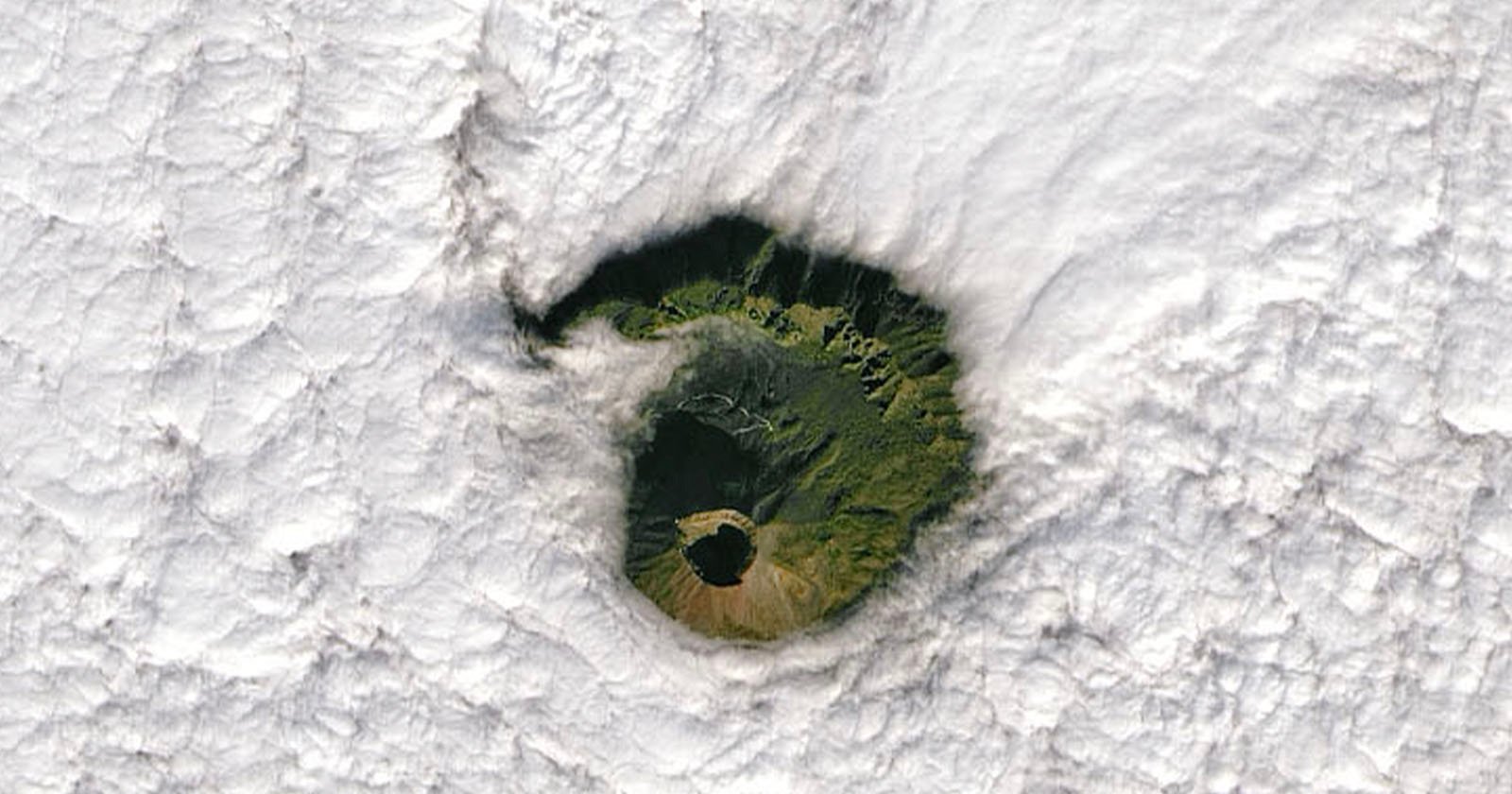 Aerial view of a volcanic island with lush greenery visible inside the crater. The surrounding area is covered by a thick layer of white clouds, creating a striking contrast with the vibrant green of the crater.