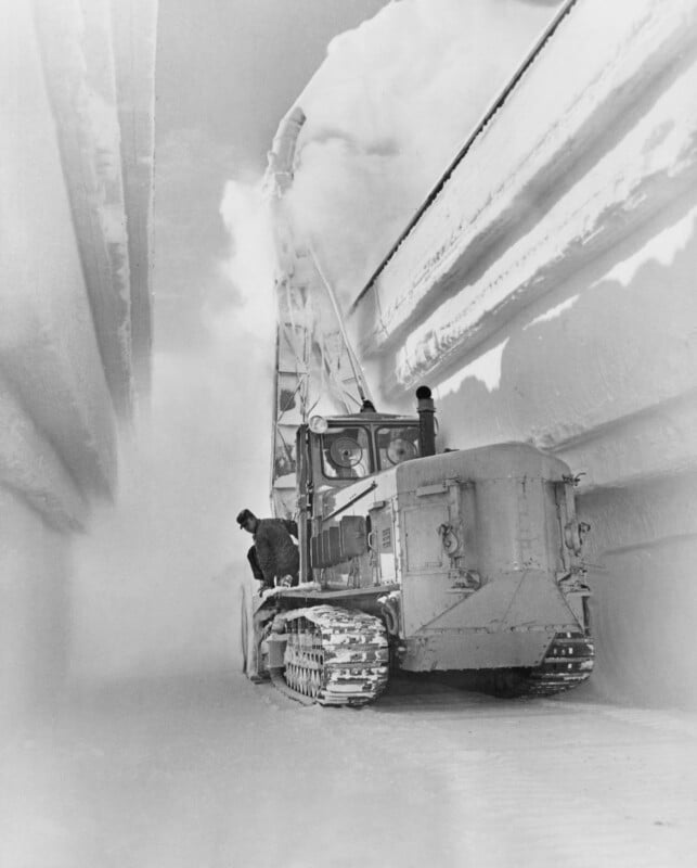 A person operates a large snow plow tractor clearing a path through deep snow. The vehicle is surrounded by towering snow walls on either side, with snow being thrown high into the air. The scene is monochromatic and wintry.