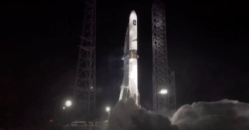 A rocket is illuminated against a dark night sky, surrounded by metal support structures. Bright spotlights and clouds of smoke or vapor enhance the dramatic scene of an impending launch at a space facility.