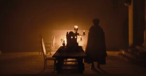 A dimly lit, atmospheric scene shows a figure in a cloak standing at one end of a long wooden table, adorned with a candelabra and goblets. A warm glow from a fireplace in the background casts shadows across the stone walls and floor.