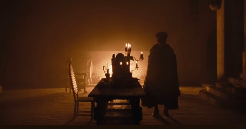 A dimly lit, atmospheric scene shows a figure in a cloak standing at one end of a long wooden table, adorned with a candelabra and goblets. A warm glow from a fireplace in the background casts shadows across the stone walls and floor.