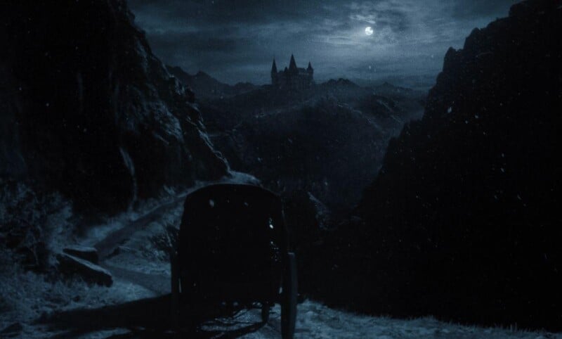 A carriage travels through a snowy, mountainous path during a dark, moonlit night. In the distance, a castle is silhouetted against the cloudy night sky, creating a mysterious and eerie atmosphere.