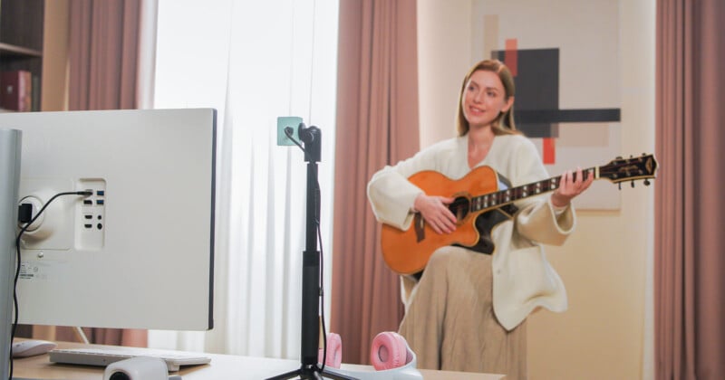 A woman plays an acoustic guitar in a softly lit room, standing near curtains and modern art. A desktop computer and headphones are visible on a table in the foreground.