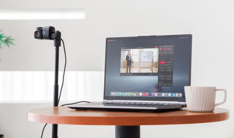 A laptop on a round table displays a video call with a person presenting at a whiteboard. A dual-lens camera on a stand is positioned beside it. A white mug sits on the table. The background is minimalistic with a hint of greenery.