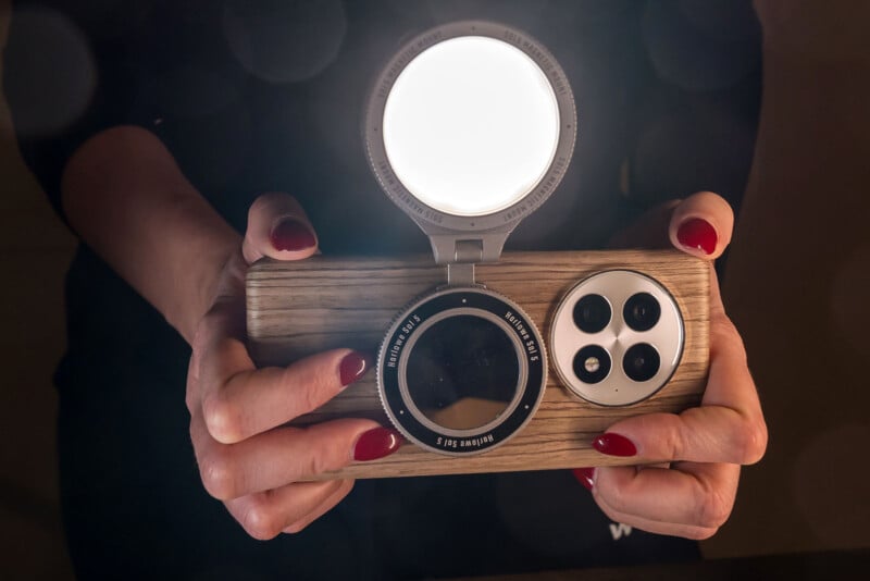 Hands holding a wooden camera accessory featuring multiple lenses and a bright circular light attachment. The person's fingernails are painted red.