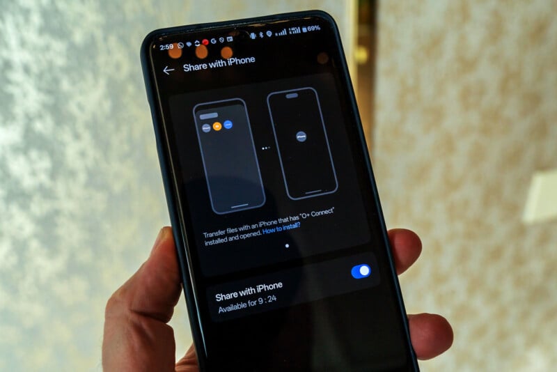 A person holding a smartphone showing a screen with instructions on sharing files with an iPhone. The screen displays two phone icons and an option to toggle "Share with iPhone," which is currently switched off.