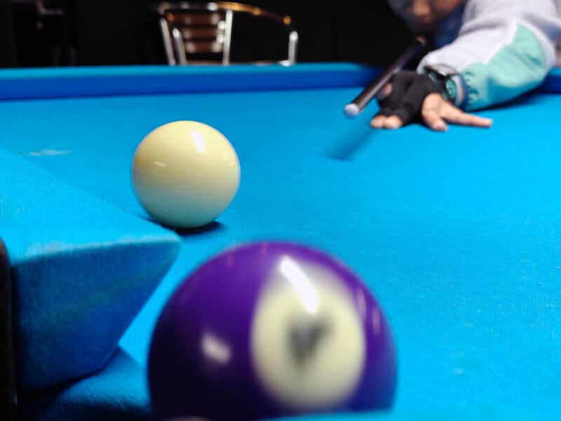 A person wearing a glove aims a cue stick at a yellow billiard ball on a bright blue pool table. A purple ball with the number 4 is blurred in the foreground. A metal chair is visible in the background.