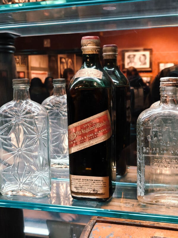 A collection of vintage glass bottles on a shelf, featuring an Old Taylor Whiskey bottle with a red and gold label in the center. The surrounding bottles are clear glass with intricate patterns, displayed on mirrored shelves.