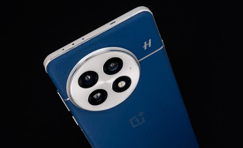 Close-up of a blue smartphone with a rectangular shape, featuring a prominent circular camera module on the back with four lenses. The phone has a metallic edge and a logo in the lower half. The background is black, highlighting the phone.