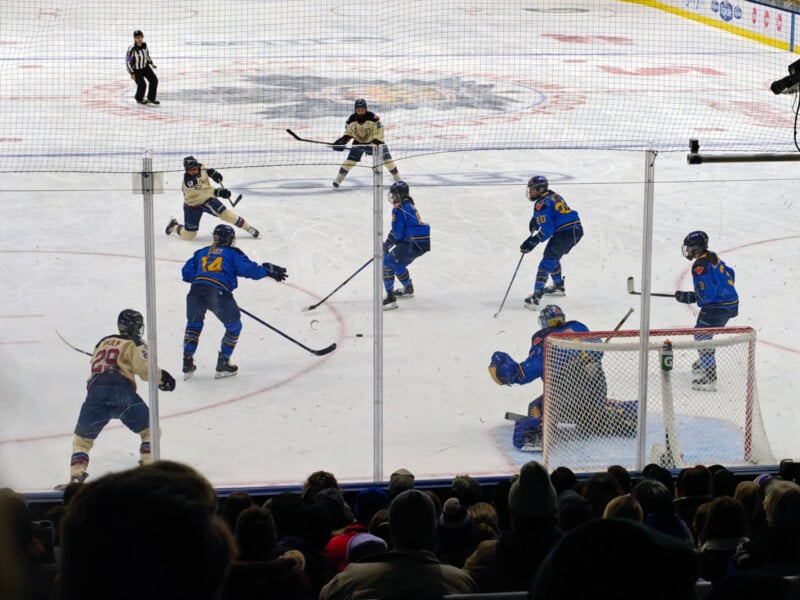 Hockey players in blue uniforms and players in white uniforms compete on the ice. One player in blue prepares to shoot the puck, while others defend. Spectators watch from the stands, and a referee stands on the rink.