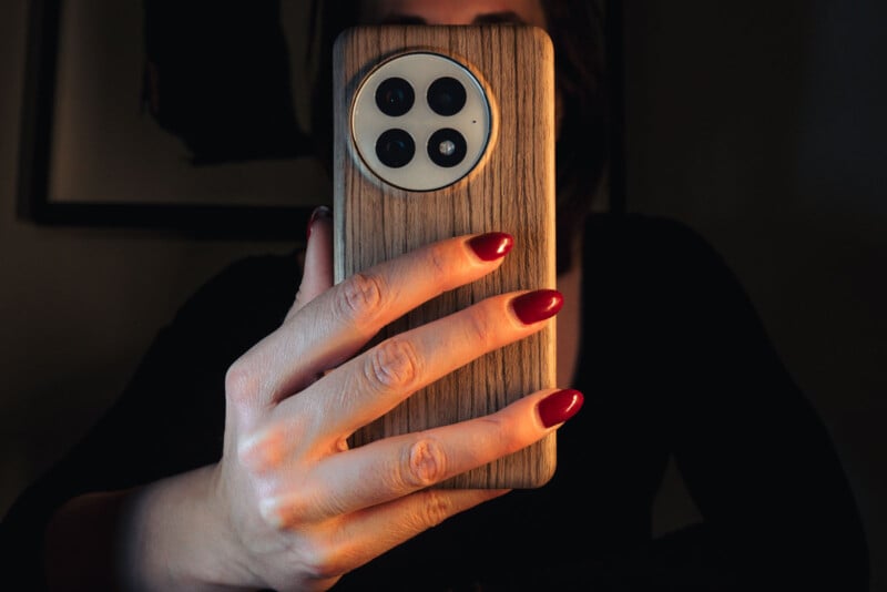 A person with red painted nails holds a smartphone with a wooden-style case, featuring three rear camera lenses. The background is dimly lit.