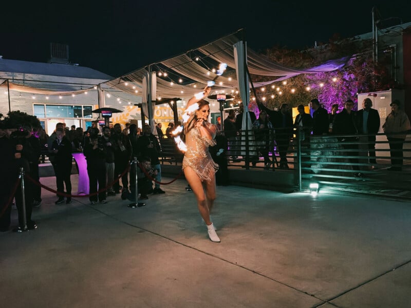 A performer in a sparkling outfit dances with flaming batons at an outdoor event. A crowd watches under string lights, with a building in the background. It's nighttime, creating a vibrant and energetic atmosphere.