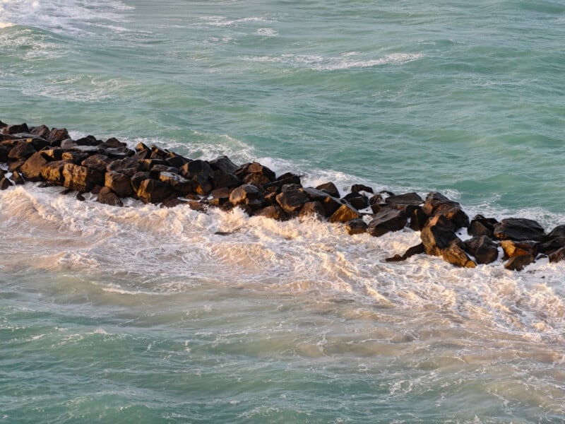 Aerial view of a rocky breakwater extending into a turquoise ocean. Waves crash against the dark rocks, creating white foam. The sunlight casts a warm glow over the scene, highlighting the contrast between the rocks and the sea.