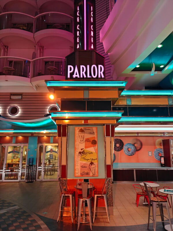 A neon-lit ice cream parlor with a retro design features a sign that reads "PARLOR" above. The interior is colorful with teal and pink accents. Tables and chairs are set up outside, and signs display various ice cream options.