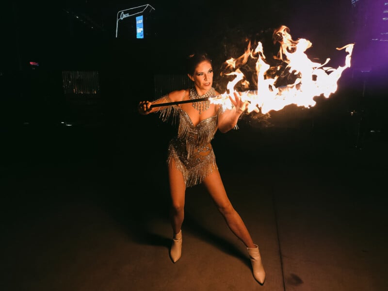 A performer wearing a sparkly outfit and white boots twirls a flaming baton outdoors at night. The background is dark with some illuminated buildings visible.